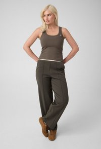 Femme blonde debout, mains derrière le dos, portant un débardeur marron, un pantalon large sombre et des chaussures marron à lacets sur un fond uni.