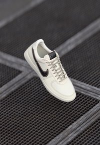 Hvide lædersneakers med brune detaljer, teksturerede snørebånd og en syet design. Har en rund tå og et logo på tungen.