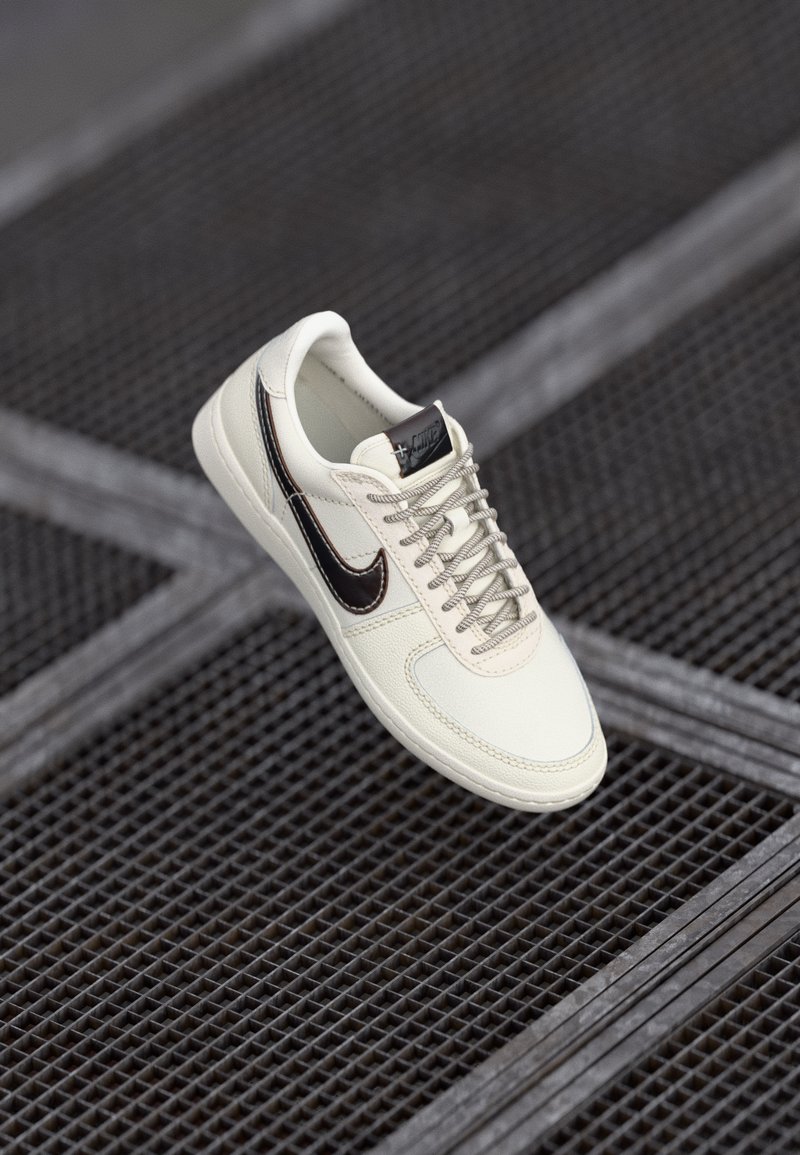 Hvide lædersneakers med brune detaljer, teksturerede snørebånd og en syet design. Har en rund tå og et logo på tungen.