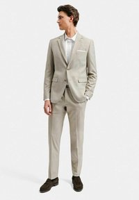 Ljusbeige kostym tillverkad av texturerad tyg, med flikiga slag, två knappar och två fickor. Matchad med en vit skjorta och bruna skor.