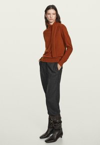 Frau in einem rostfarbenen Pullover mit angenähtem Schal, dunkelgrauen, lockeren Hosen und schwarzen, lässigen Stiefeln, die mit den Händen in den Taschen steht.