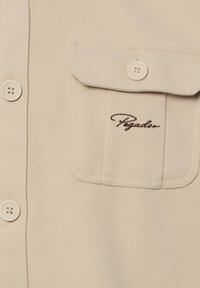 Beige Hemd mit Knopfleiste aus strukturiertem Stoff, mit einer Brusttasche, auf der das braune "Pegador"-Logo gestickt ist. Vier Knöpfe sichtbar.