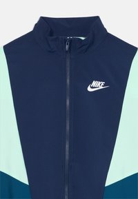 Tamno plava sportska jakna s patentnim zatvaračem, s akcentima u tirkiznoj i tamno tirkiznoj boji, na lijevoj strani prsnog koša s bijelim Nike logotipom.