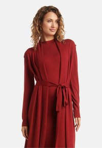 Long, red, knitted dress with a wrap design, featuring a tie waist and long sleeves. The fabric appears soft with a smooth texture.