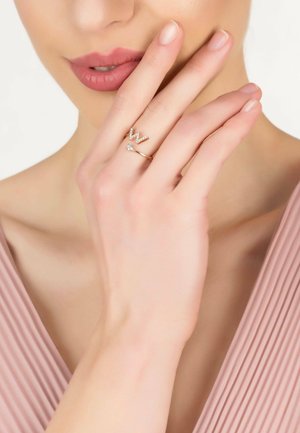 Gold rings with clear gemstones; one features a "W" design. Smooth skin, light pink blouse. Hand positioned near face.