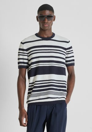 Hombre con gafas de sol negras, suéter blanco de manga corta con rayas horizontales azul marino y pantalones azul marino, de pie frente a un fondo liso.