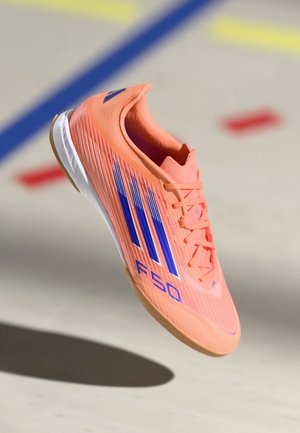 Chaussure de football en salle orange vif avec des rayures bleues et une étiquette "F50", présentée sur un fond flou de terrain.