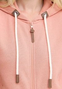 Haut à capuche rose avec une fermeture éclair, présentant un tissu texturé, un cordon blanc avec des pointes marron et des accents marron au niveau du cou.