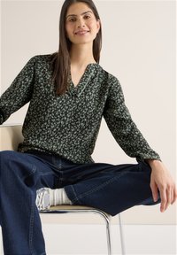 Woman sitting on chair with legs apart wearing a green patterned blouse, dark blue jeans, white socks, and white sneakers.