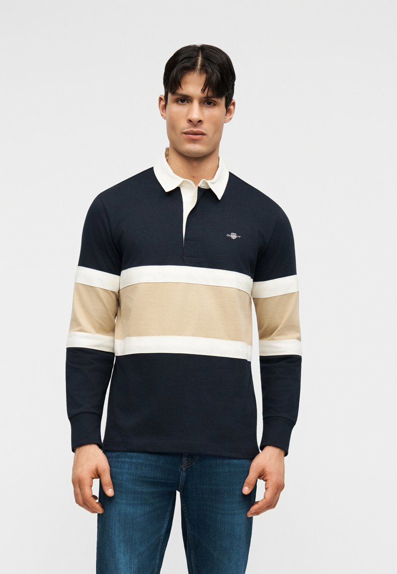 Homme portant un maillot de rugby à manches longues rayé bleu marine, beige et blanc avec un col blanc, et un jean bleu, debout devant un fond uni.