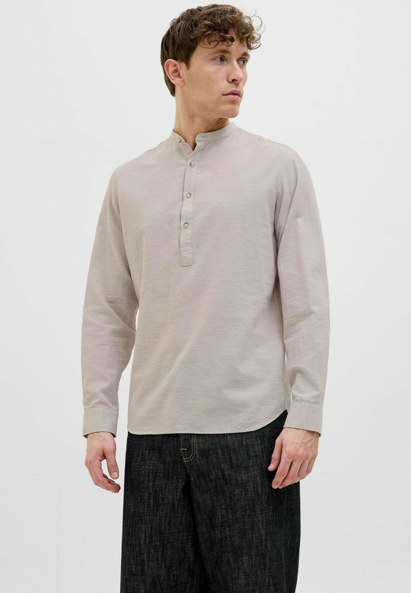 Lys beige langærmet skjorte med stående krave, tre-knaps lukning og buet kant. Parret med mørke denim-bukser.