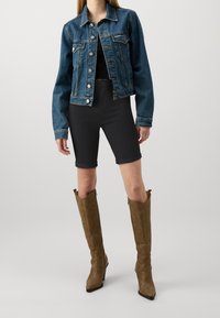 Chaqueta de mezclilla en azul oscuro con botones plateados, combinada con pantalones cortos negros de tiro alto y botas de cuero marrón hasta la rodilla con punta afilada.