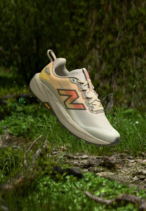 Laufschuh mit orangefarbenen und gelben Akzenten, der über einem grasbewachsenen Waldboden mit Erde und kleinen Pflanzen schwebt.