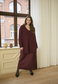 Maglione in maglia oversize bordeaux con scollo a V, abbinato a una gonna maxi in satin coordinata. Le scarpe flat con dettaglio a fiocco nero completano il look.