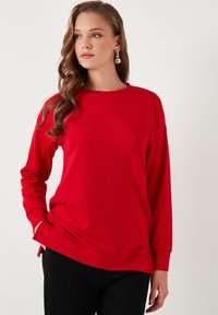 Maglietta rossa a maniche lunghe con scollatura rotonda, realizzata in tessuto morbido. Presenta spacchi laterali e una vestibilità rilassata. Abbinata a pantaloni neri.