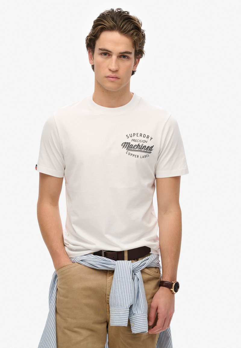 Witte katoenen t-shirt met "Superdry Machined Copper Label" opdruk, korte mouwen, gecombineerd met lichtbruine broek en een blauw gestreept shirt om de taille geknoopt.