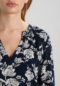 Květinová blůza s tmavě námořnickým pozadím a bílými botanickými vzory, výstřih do V a měkkou, strukturovanou tkaninou.