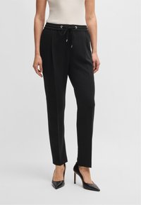 Pantaloni neri con vita regolabile, vestibilità affusolata, tessuto liscio e occhielli metallici. Abbinati a scarpe con tacco alto nere.