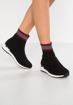 Ben iført sorte sokkelignende sneakers med hvide såler og røde og grå stribede manchetter på en glat, lys gulv.