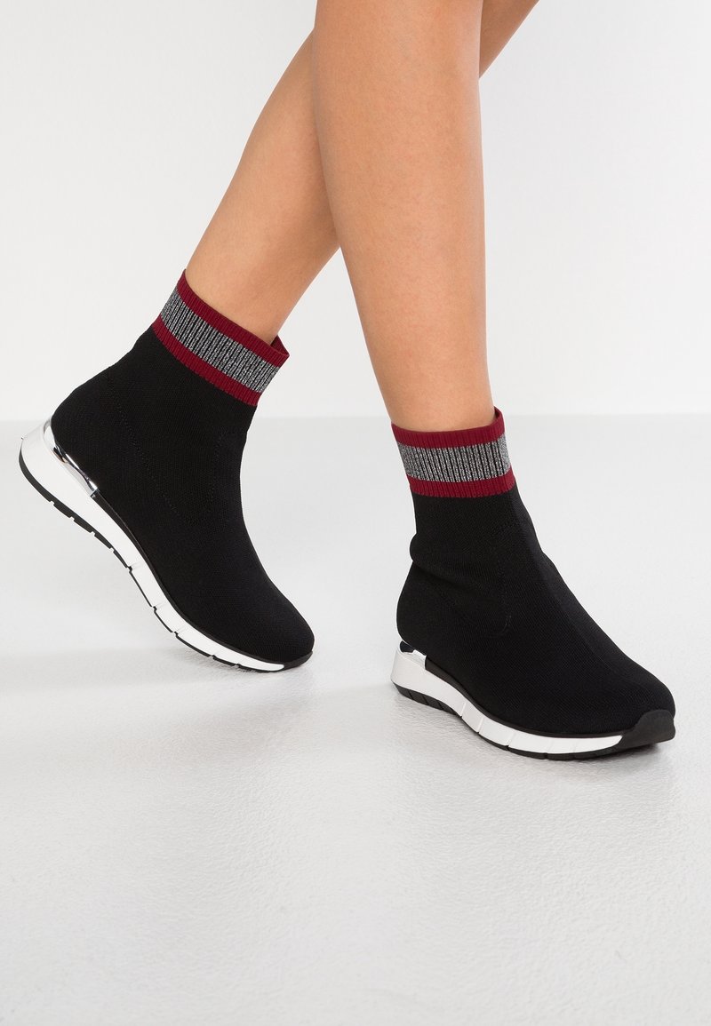 Jambes portant des baskets style chaussettes noires avec semelles blanches et bords rayés rouges et gris sur un sol lisse et clair.