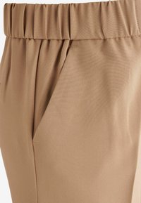 Pantalon beige avec une taille élastique, dotée de plis à l'avant et d'un tissu doux et texturé. Poches latérales comprises pour la fonctionnalité.