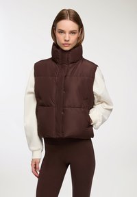 Gilet imbottito marrone con colletto alto, dotato di chiusura a scatto frontale e cuciture orizzontali. Indossato sopra una maglietta bianca a maniche lunghe e leggings marroni.