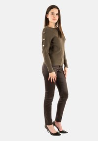 Maglione verde oliva con scollatura rotonda e bottoni decorativi sulla manica sinistra, abbinato a pantaloni marroni aderenti e tacchi alti neri.