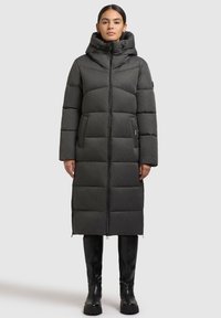 Harmaa pitkä puhvitakki hupulla, jossa on vetoketjukiinnitys, vaakasuorat tikatut osat ja sivutaskut. Tyylitelty mustilla polvipitkillä saappailla.