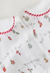 Tissu blanc avec des imprimés festifs d'ornements de Noël, de cannes de bonbon et de pain d'épice, avec des boutons-pression rouges et un col festonné.