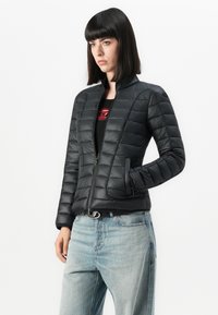 Svart pufferjakke med glidelås i front, med en figursydd design, horisontal søm og sidelommer. Brukt med lyseblå jeans.