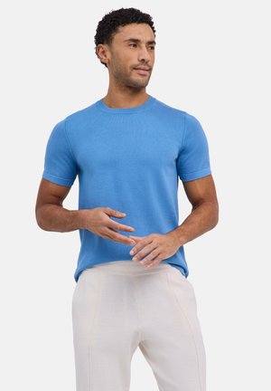 Mann trägt ein blaues Kurzarmhemd und hellbeige Hose, steht mit locker verschränkten Händen und schaut zur Seite.