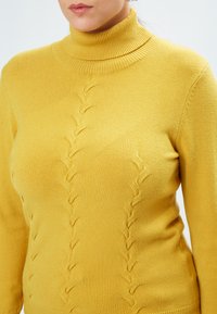 Suéter amarillo de cuello alto hecho de una tela suave, con un collar acanalado y un detallado texturizado en forma de hojas verticales en la parte delantera.