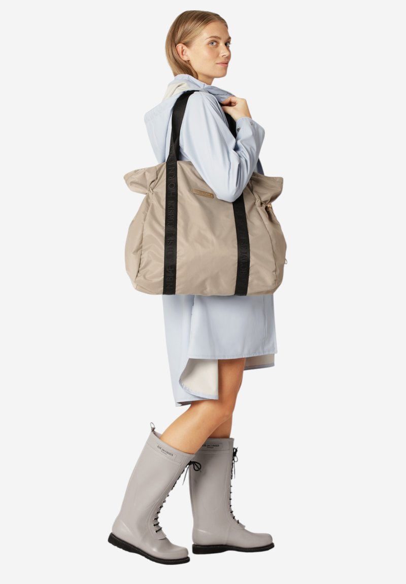 Beżowa, oversize torba nylonowa z czarnymi uchwytami. Modelka ma na sobie jasnoniebieską kurtkę przeciwdeszczową oraz szare gumowe buty z sznurówkami. Tło jest białe.
