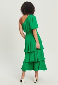 Zelené šaty na jedno rameno s objemovým rukávem, vrstvenými volánky a splývavým střihem; vyrobeny z mačkané, lehké látky.
