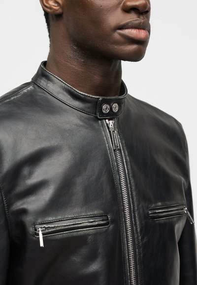 Κοντινό πλάνο ενός ατόμου που φοράει μαύρο δερμάτινο μπουφάν με φερμουάρ μπροστά, δύο τσέπες στο στήθος με φερμουάρ και γιακά με κουμπί πίεσης.