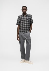 Νέος άνδρας που φοράει γκρι καρό κοντομάνικο πουκάμισο, γκρι παντελόνι cargo και μπεζ αθλητικά παπούτσια, όρθιος μπροστά σε απλό φόντο.