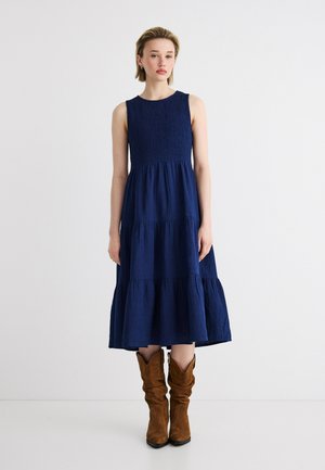 Vrouw met kort blond haar, staand, gekleed in een navyblauwe mouwloze midi-jurk met meerdere lagen en bruine suède laarzen met een slouchy pasvorm.