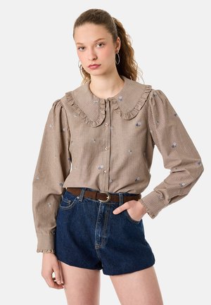 Ung kvinde med creol øreringe iført en beige stribet bluse med flæsekant og blå blomsterdetaljer, kombineret med denimshorts og et brunt bælte.
