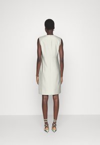 Robe sans manches ajustée en tissu gris clair, avec une silhouette cintrée, une fermeture éclair au dos et un ourlet à hauteur du genou, associée à des talons verts à fines brides.