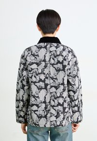 Persona con capelli corti scuri che indossa una giacca floreale nera e grigia e jeans blu, fotografata da dietro su uno sfondo bianco.