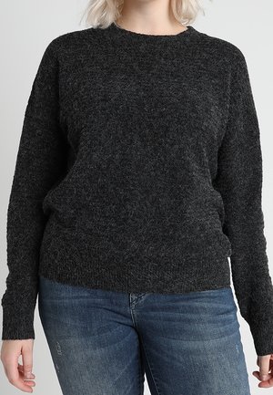 Kobieta w ciemnoszarym, puszystym swetrze i niebieskich dżinsach, stojąca na neutralnym tle z rękami swobodnie opuszczonymi wzdłuż ciała.