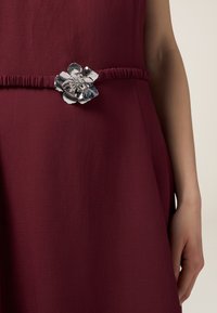 Rochie burgundy cu un finisaj texturat; prezintă o talie strânsă și o curea florală argintie cu detalii intricate.