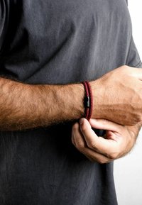 Červený zapletaný náramok s čiernym uzáverom, nosený na zápästí. Detailný záber na textúru a dizajn, zdôrazňujúci zložitý vzor tkania.