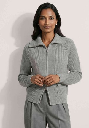 Femme portant un pull zippé côtelé gris clair avec un large col et un pantalon plissé gris assorti, debout les mains jointes.