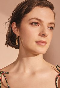 Guldörhängen i form av hoops med en böjd design och en fäst länkdetalj. Håret är kort och fuktigt, mot en mjuk rosa bakgrund.