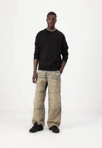 Svart hettegenser, beige cargobukser med lommer og stripete design, og svarte sneakers. Modellen poserer sentrert mot en hvit bakgrunn.