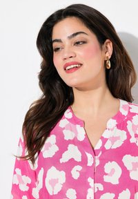Rosa Hemd mit weißen Leopardenmuster, kurze Ärmel und Kragen. Der Stoff wirkt leicht und glatt.