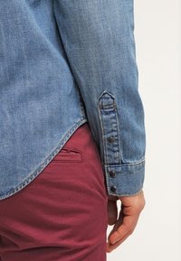 Personne portant une chemise en jean bleue avec des poignets boutonnés et un pantalon rouge, main détendue le long du corps sur un fond uni.