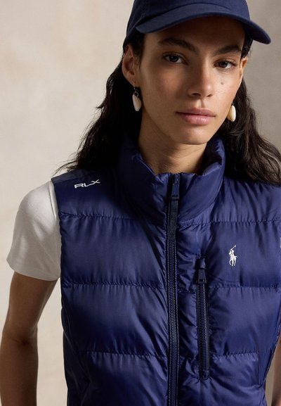 Γυναίκα που φοράει σκούρο μπλε καπιτονέ γιλέκο με φερμουάρ και λογότυπο, λευκό κοντομάνικο πουκάμισο, σκούφο σε ναυτικό μπλε και σκουλαρίκια σε σχήμα κοχυλιού, κοιτάζοντας προς τα εμπρός.