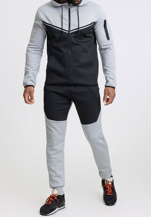 Survêtement noir et gris avec une veste à capuche zippée. Présente un design en chevron, plusieurs poches et des textures contrastantes. Inclut des baskets noires avec des lacets orange.